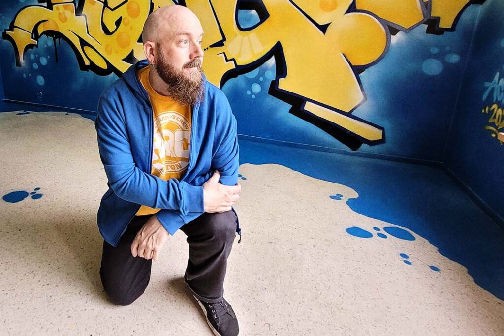 Bald and bearded person inside a room that is painted with graffiti.
