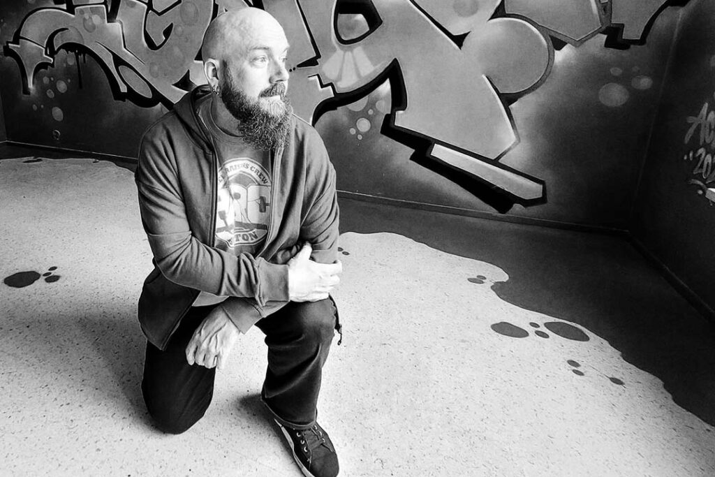 Bald and bearded person inside a room that is painted with graffiti.
